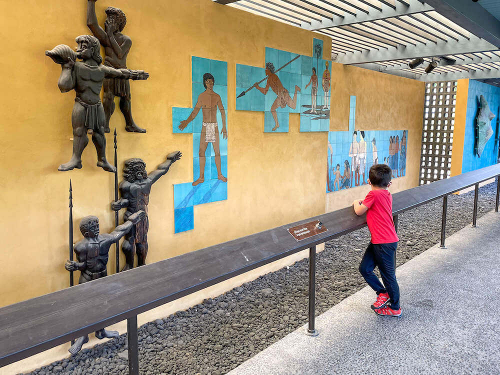 Image of a boy standing on a railing with Hawaiian artwork on a yellow wall in front of him.