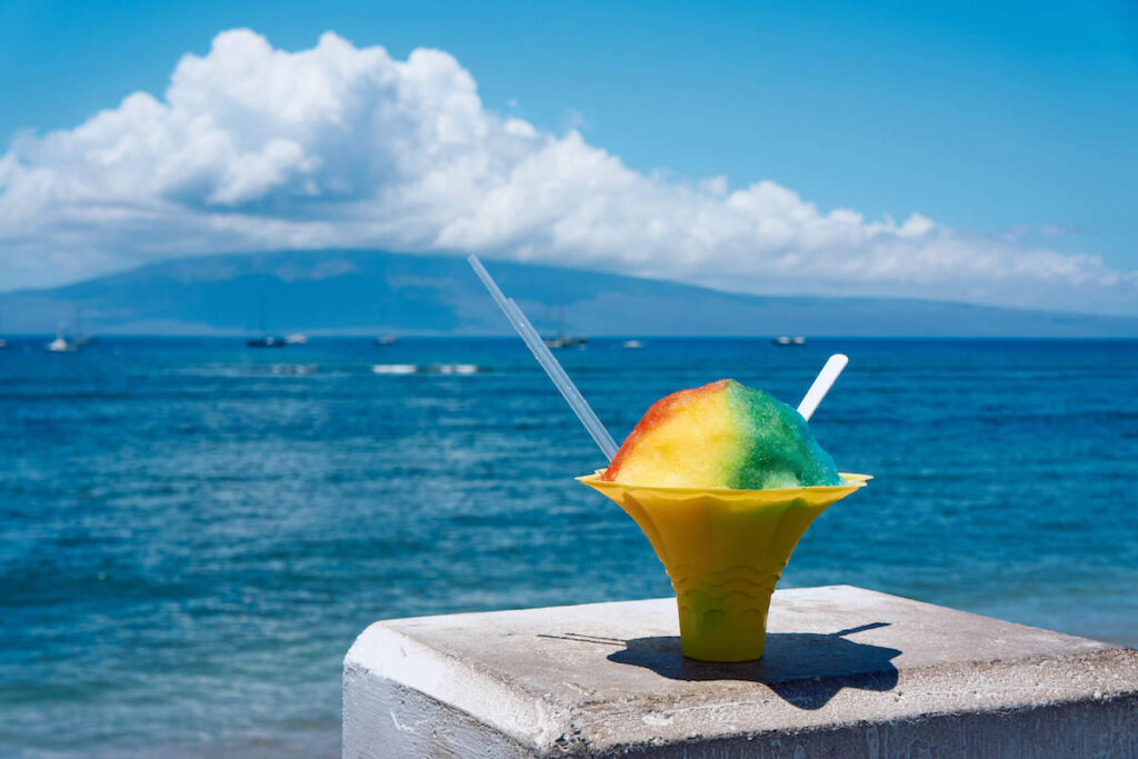 Find out where to get the best shave ice in Maui Hawaii by top Hawaii blog Hawaii Travel with Kids. Image of a rainbow shave ice in a yellow cup sitting on a concrete pillar with the Pacific Ocean in the background.