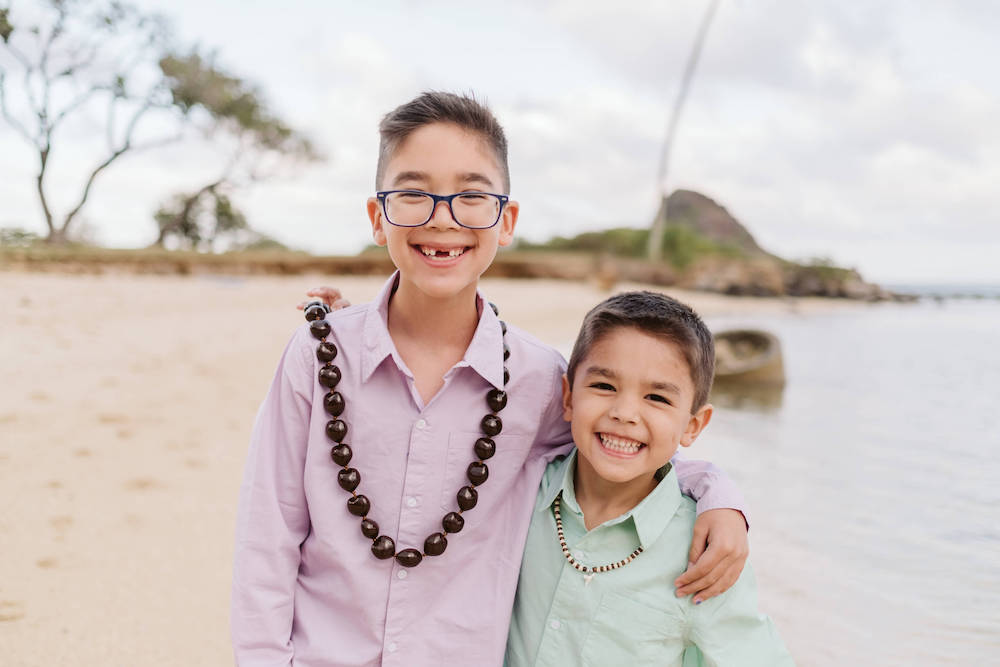 Image of two boys wearing button down shirts with their arms around each other smiling at the camera on a beach in Oahu Hawaii