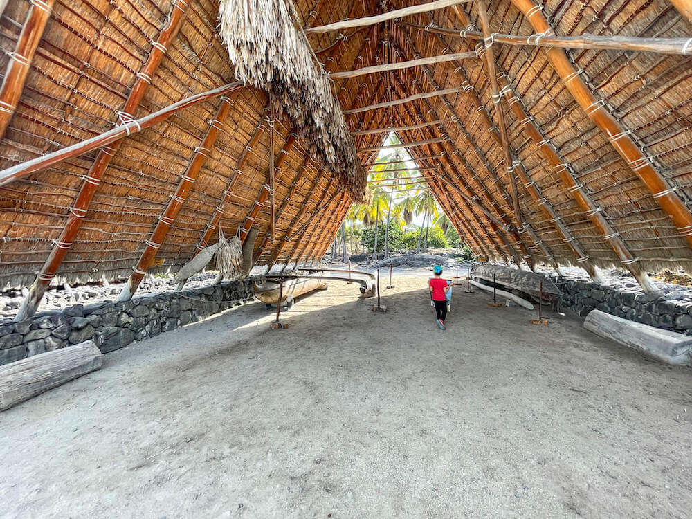 Image of a boy walking through a huge thatched Hawaiian shelter with a canoe inside.