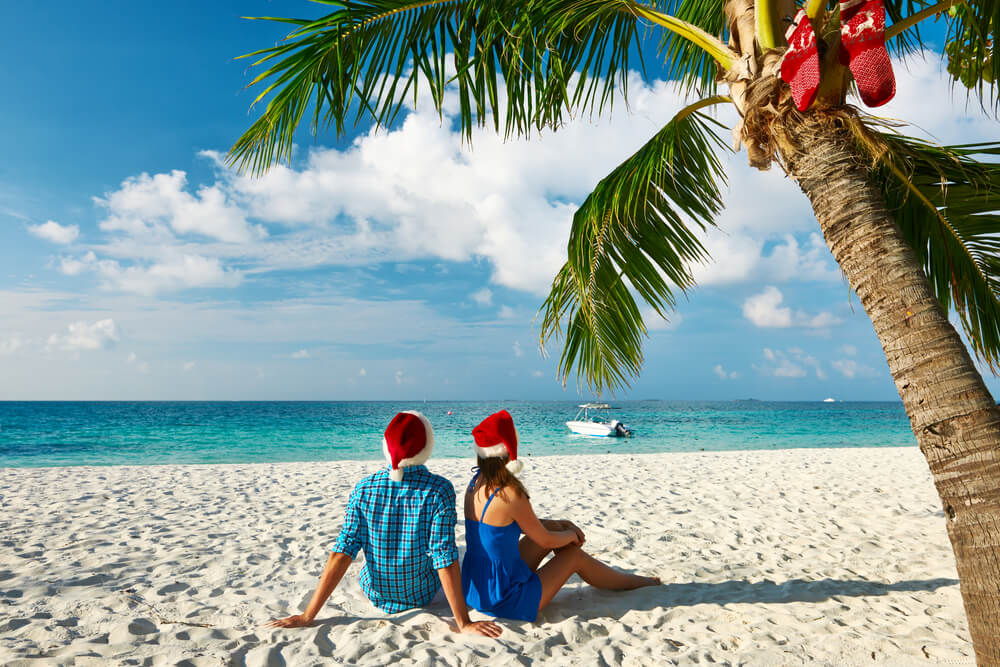 Image of two people sitting on a beach wearing Santa hats and a palm tree with stockings.