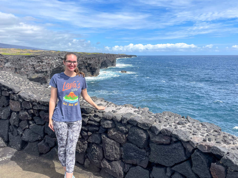 Image of a woman standing next to a lava rock wall at the edge of a cliff leading to the ocean at Hawaii Volcanoes National Park