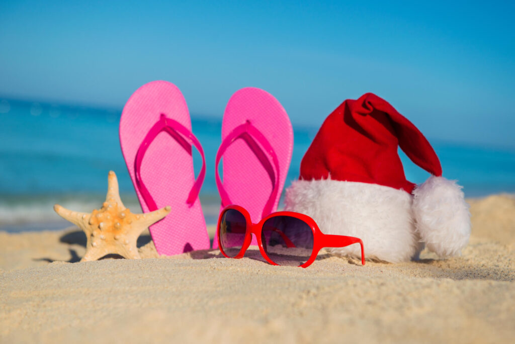 Image of pink flip flops, red sunglasses, a starfish, and a Santa hat on a beach.