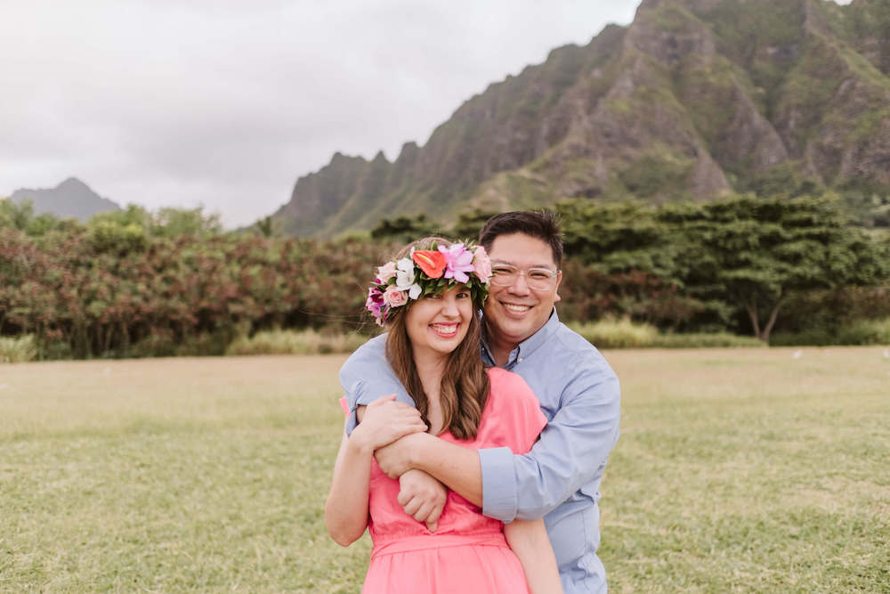 Image of a man wearing a blue shirt and a woman wearing a flower crown and pink dress posing for a photo at Kualoa Beach Park on Oahu.