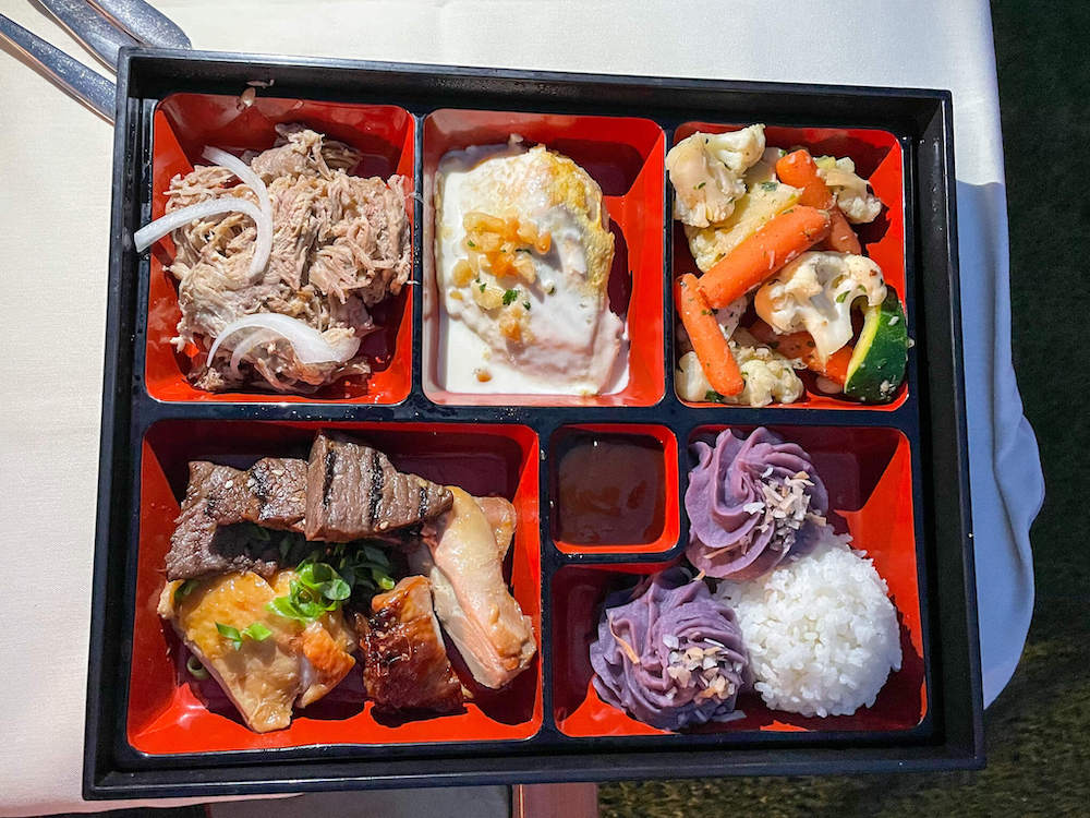 Image of a bento box filled with kalua pork, fish, steamed veggies, teriyaki steak, Hawaiian chicken, rice, and purple mashed potatoes.