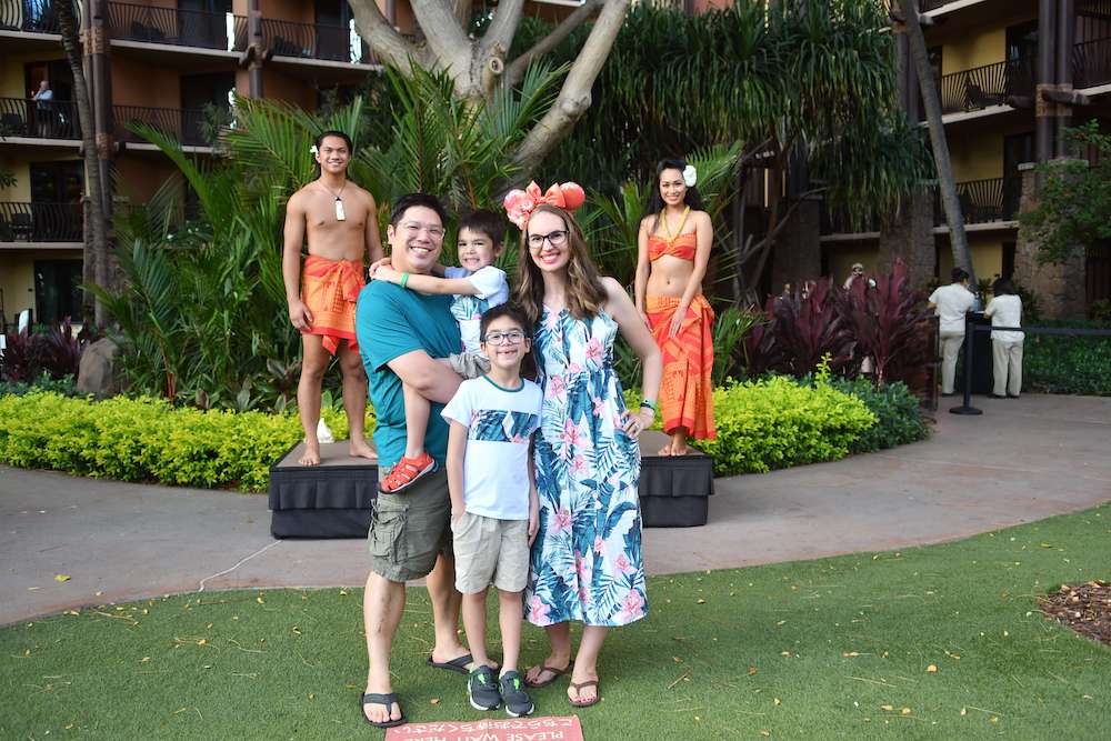 Image of a family wearing coordinating Aloha outfits posing for a photo with Disney Aulani luau dancers.