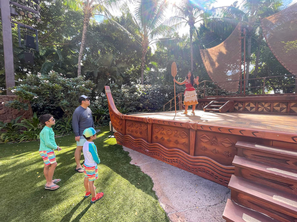 Image of Moana standing on a large stage that looks like her canoe while she talks with a man and two boys.