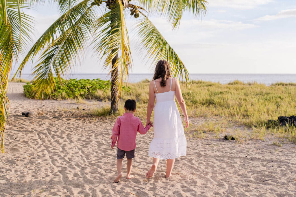 Image of a mom wearing a white dress and 5yo boy wearing a red shirt facing away from the camera on a Kona beach photo shoot in Hawaii.