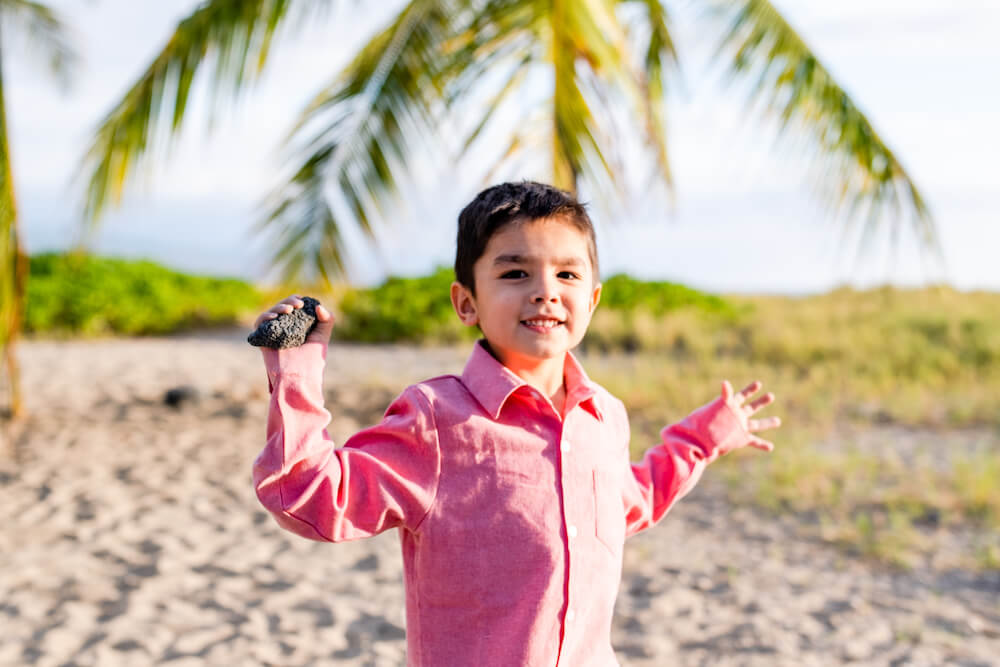 Image of a boy wearing a red button down shirt holding a lava rock on a Kona beach at sunset.
