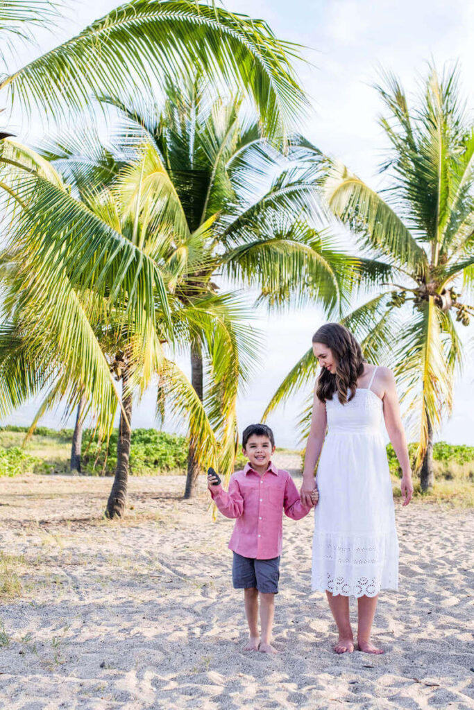 Flytographer has many Big Island photographers like Amanda who can capture beautiful Kona photoshoots like this one. Image of a mom wearing a white dress and a 5yo boy wearing a red shirt walking on the beach in Kona.