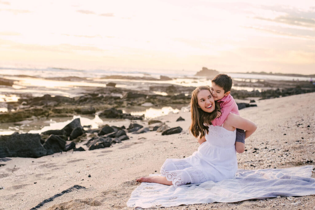 Image of a mom and son snuggling on a blanket on a beach during sunset on Kona.