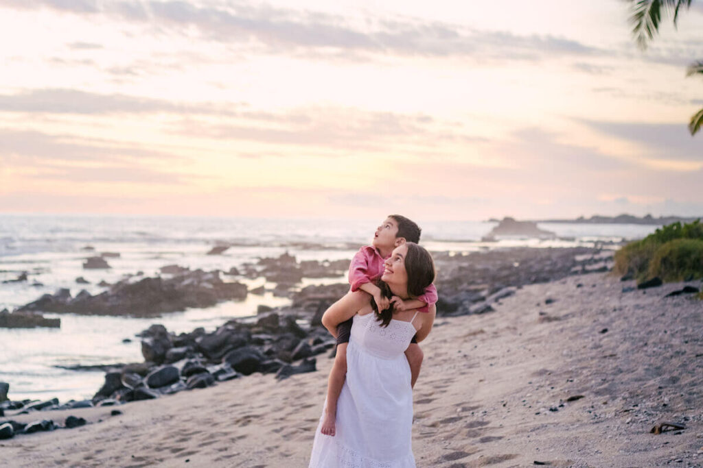 Image of a mom carrying her son on her back on a beach in Hawaii while they both look up toward the sky.