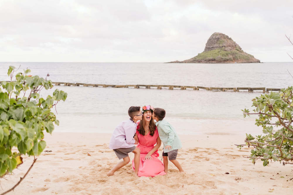 Image of a mom in a pink dress sitting on the beach with two little boys kissing her cheeks with Mokoli'i island in the background.