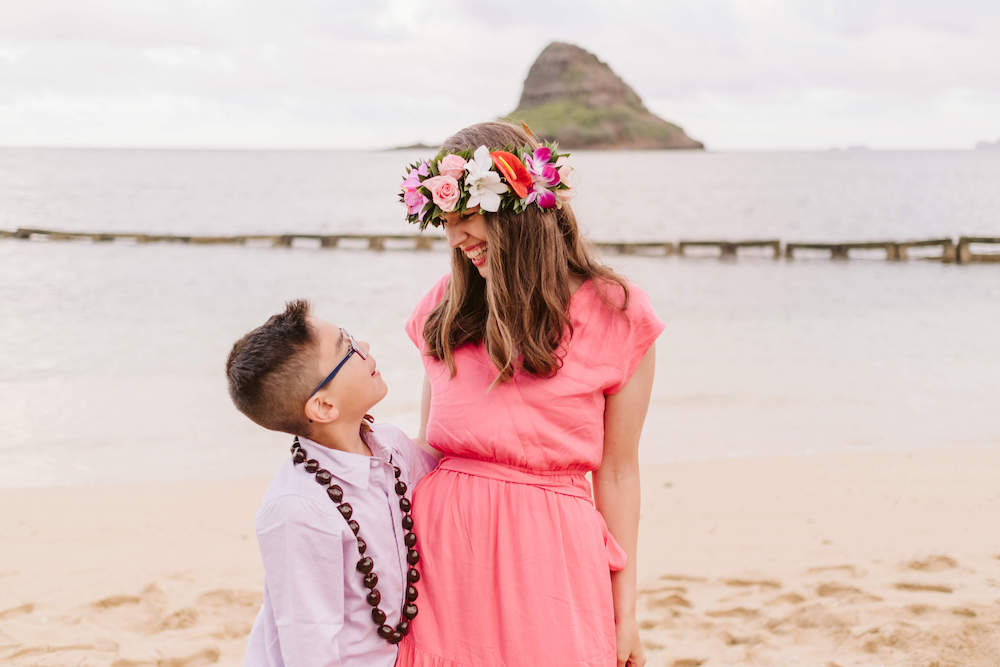 Image of a woman wearing a flower crown and pink dress looking at a boy in a purple shirt and kukui nut lei at a beach in Hawaii.