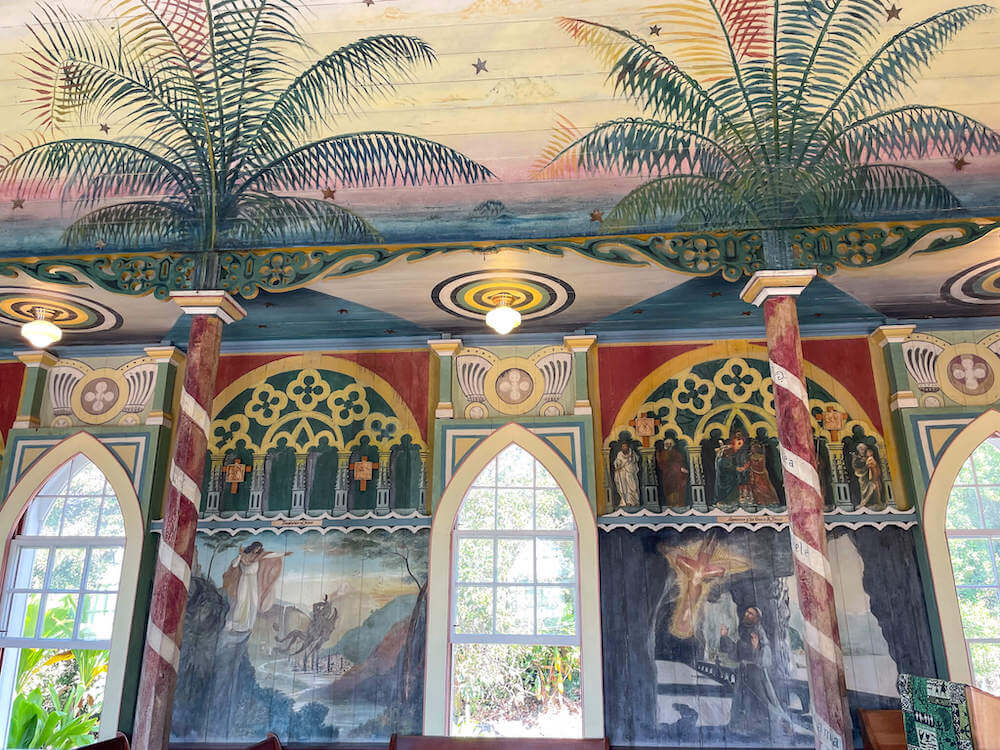 Image of a colorful church wall featuring religious paintings and palm trees that go up to the ceiling.