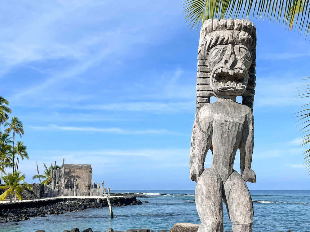 The Place of Refuge is a top Hawaii national park. Image of a tiki with a Hawaiian hut and the ocean in the background.