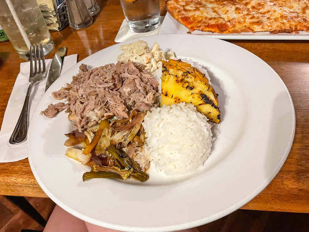 Image of kalua pork, white rice, macaroni salad, veggies, and grilled pineapple on a white plate with pizze in the background.