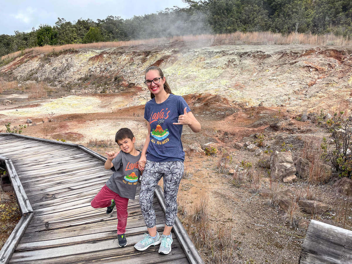 Find out what you need to know about visiting Hawaii Volcanoes National Park with kids by top Hawaii blog Hawaii Travel with Kids. Image of a mom and boy standing on a wooden boardwalk surrounded by sulphur banks in Hawaii.
