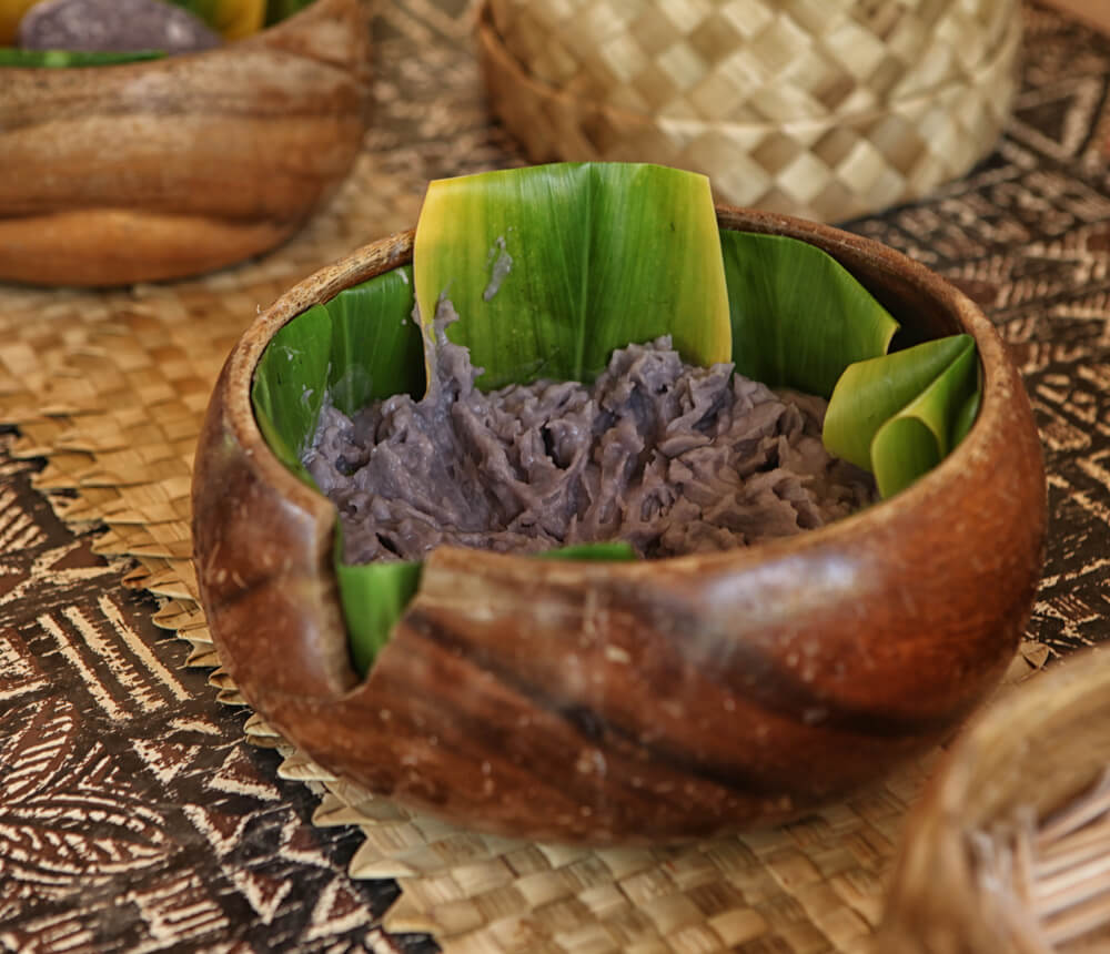 Image of a wooden bowl with ti leaves and mashed up taro root.