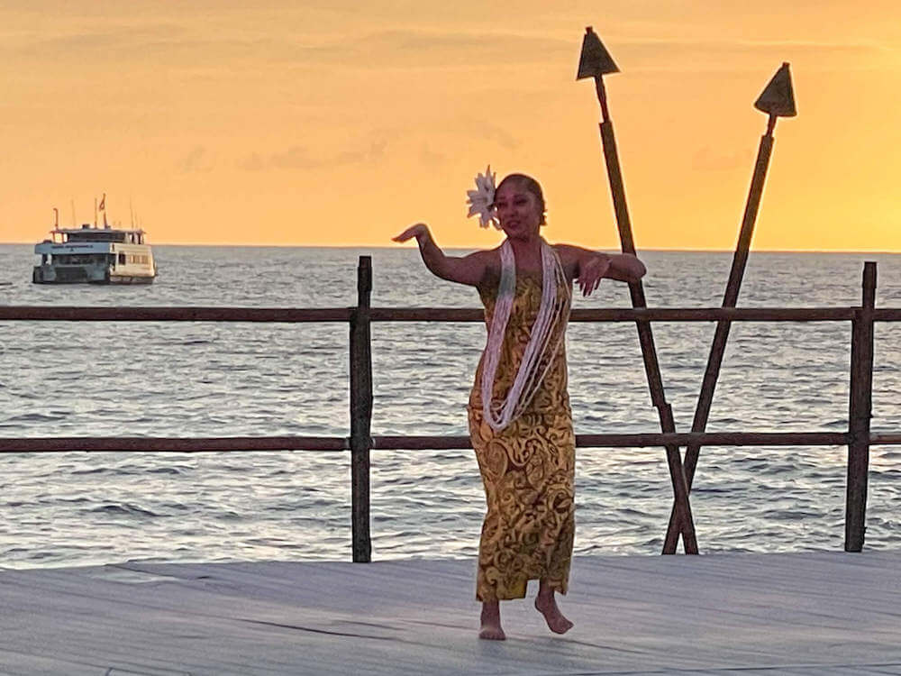 Image of a woman wearing a yellow and black printed dress dancing on stage in front of the ocean, a boat, and two tiki torches.