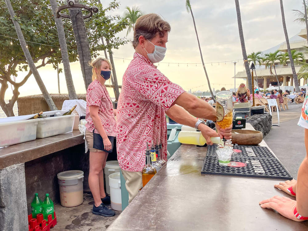 Image of a man wearing a red Aloha shirt and a white mask pouring cocktails at the Royal Kona Resort luau.