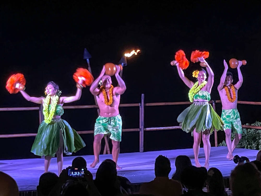 Image of women wearing ti leaf skirts and men wearing green shorts dancing with red feather rattles and gourd drums.