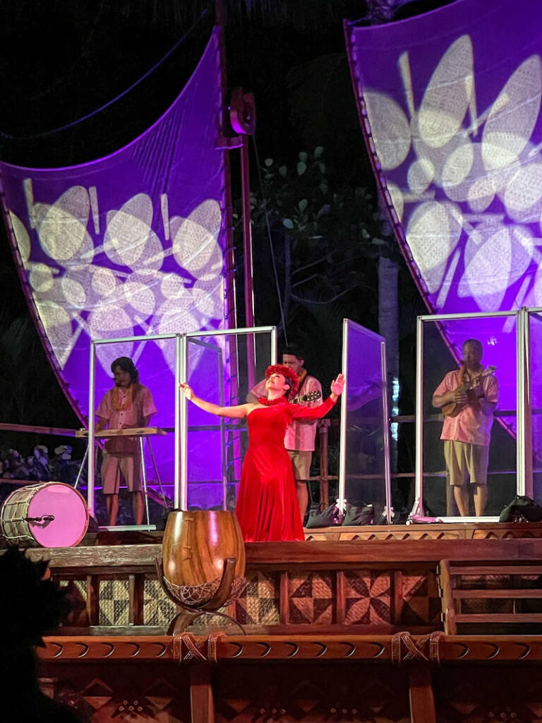 Image of a woman wearing a long red dress hula dancing at the Ka Wa'a Luau at Aulani.