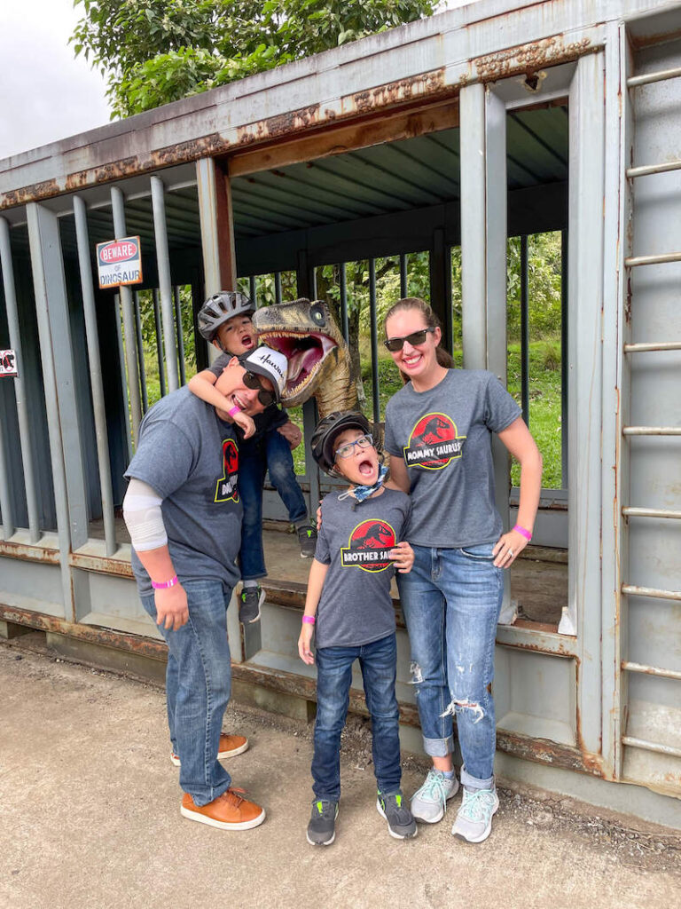 Image of a family of four wearing Jurassic Park shirts and posing with a fake dinosaur in a cage at a Jurassic World movie site.