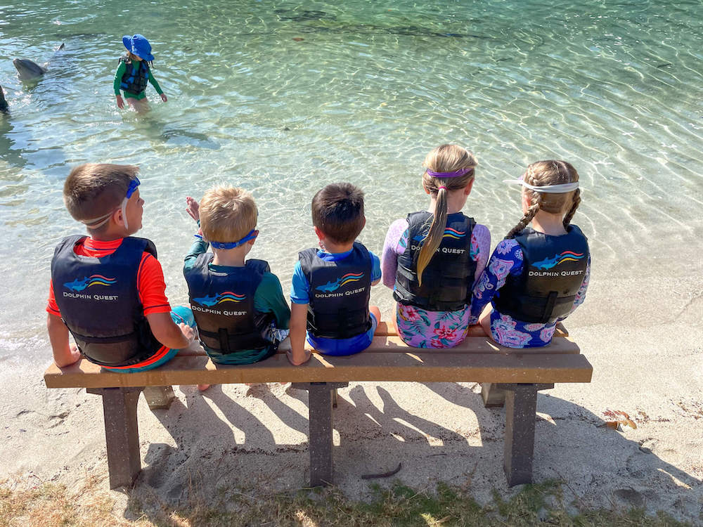 Image of kids wearing life jackets sitting on a bench facing the water.