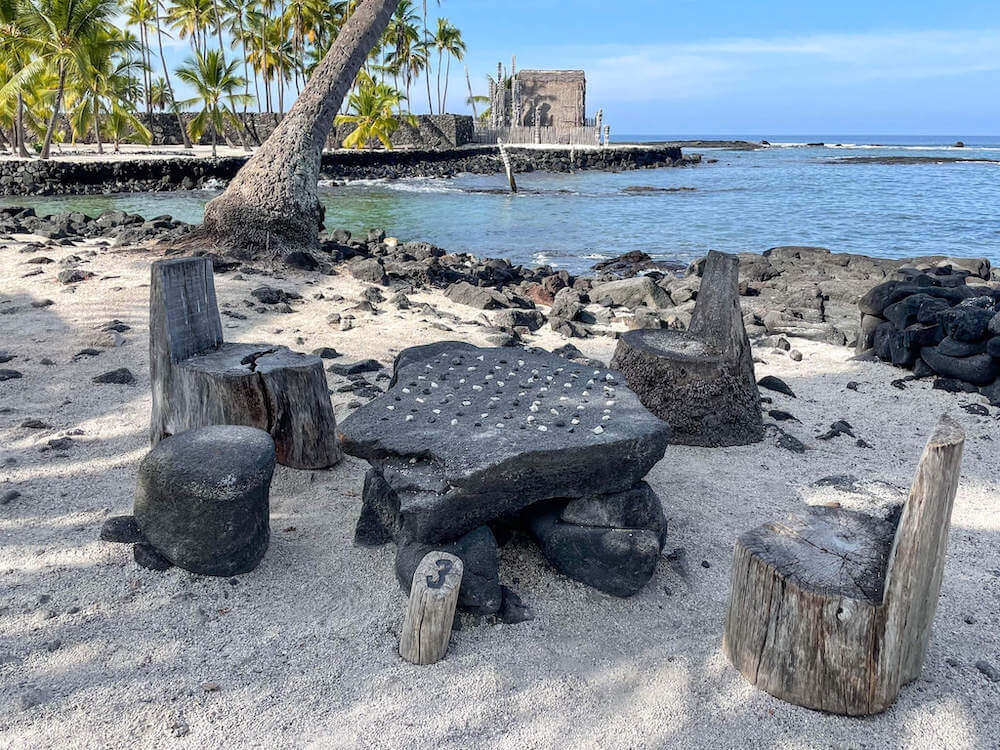 Image of a stone table with little white and black stones on it surrounded by chairs made of out wooden stumps at puuhonua o honaunau.