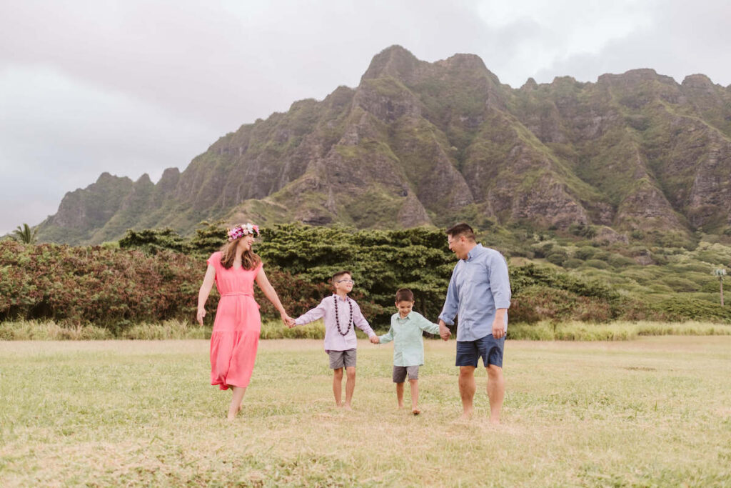 Image of a family holding hands and walking in a grassy field with Kualoa Mountains in the background.