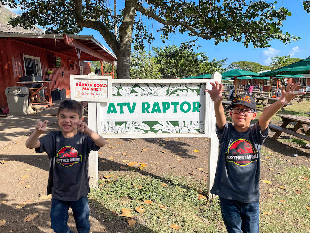 Image of two boys pretending to be dinosaurs while wearing Jurassic Park shirts and standing next to the Kualoa Ranch ATV Raptor sign.