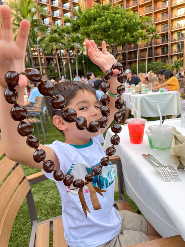 Image of a boy holding up brown kukui nut lei at the Disney Aulani luau on Oahu Hawaii.