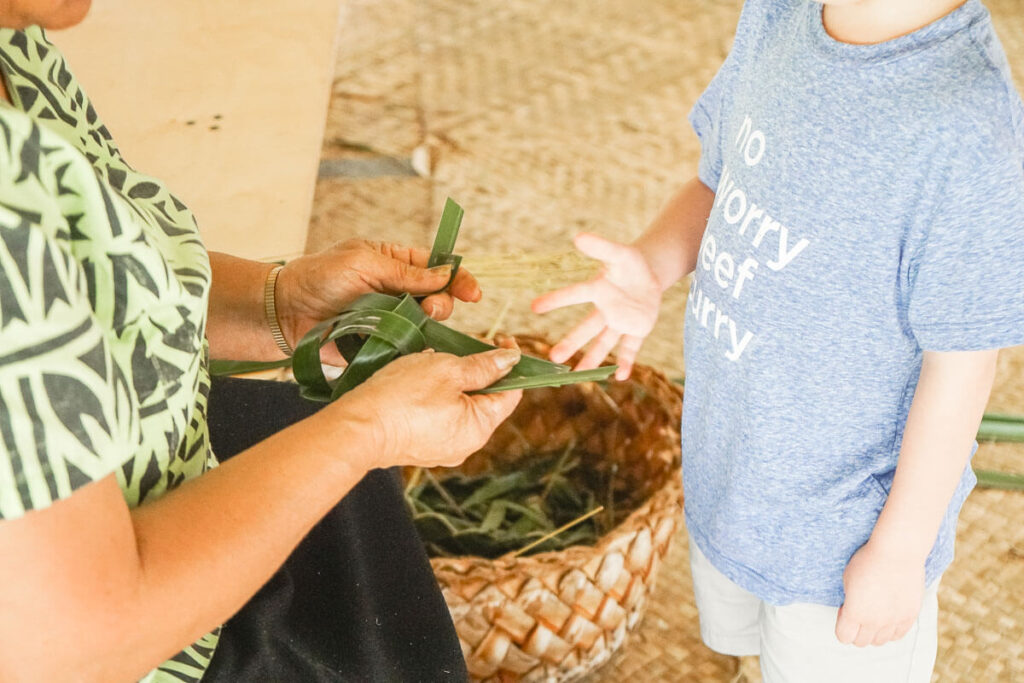 There are tons of hands on Oahu activities for kids at the Polynesian Cultural Center. Image of a woman weaving a lauhala fish for a boy.