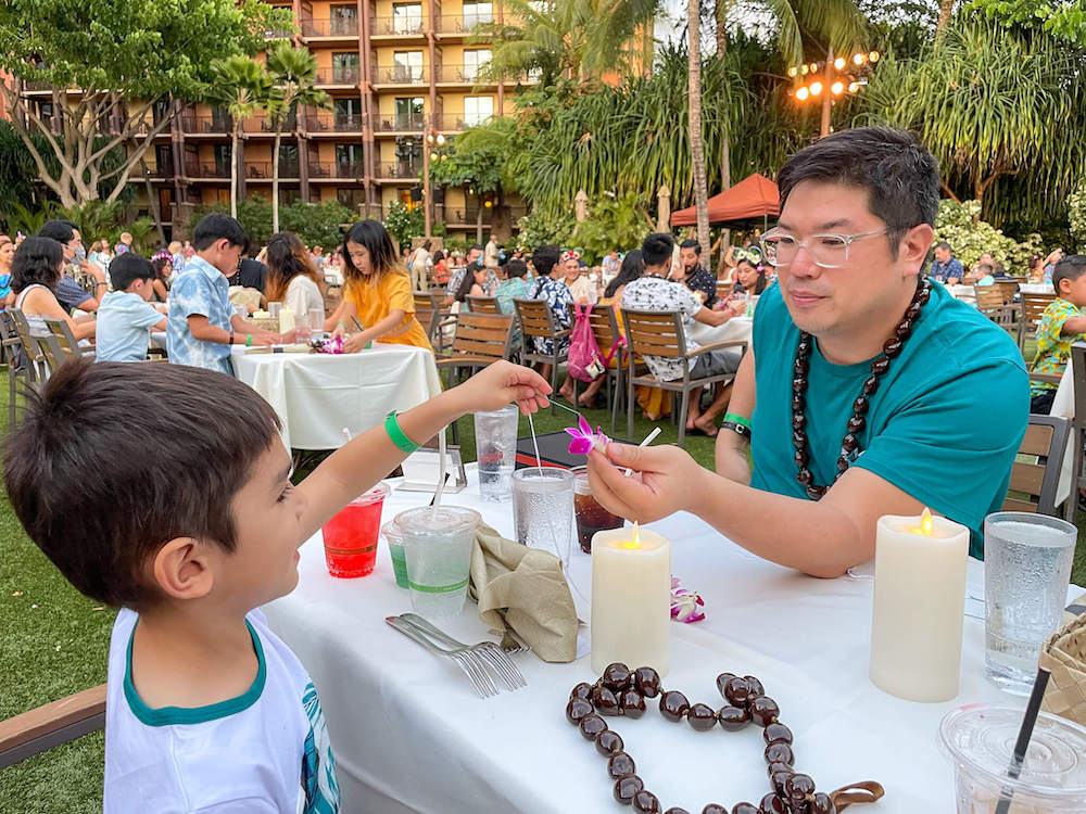 Image of a man and boy stringing a purple orchid lei at a table at the Disney Aulani luau in Oahu Hawaii.