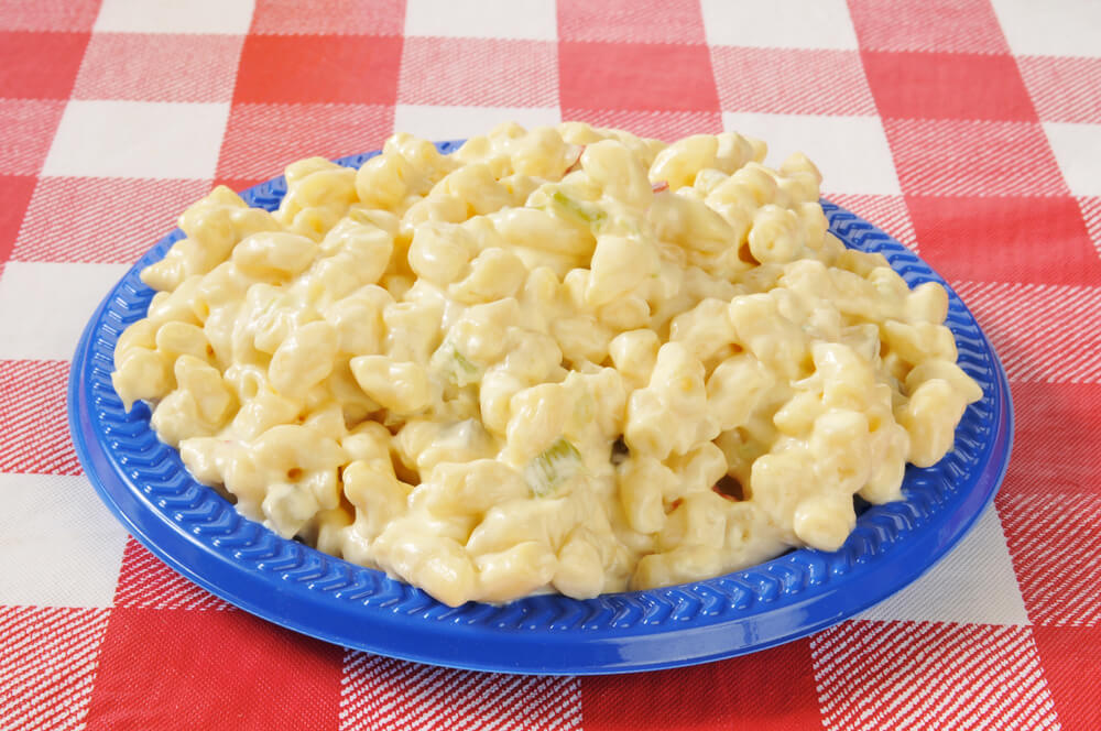 Image of a blue bowl filled with macaroni salad on a red and white checkered tablecloth.