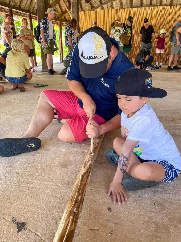You can learn how to make fire in the Samoan Village at the Polynesian Cultural Center in Hawaii. Image of a dad and son using sticks to make a fire.