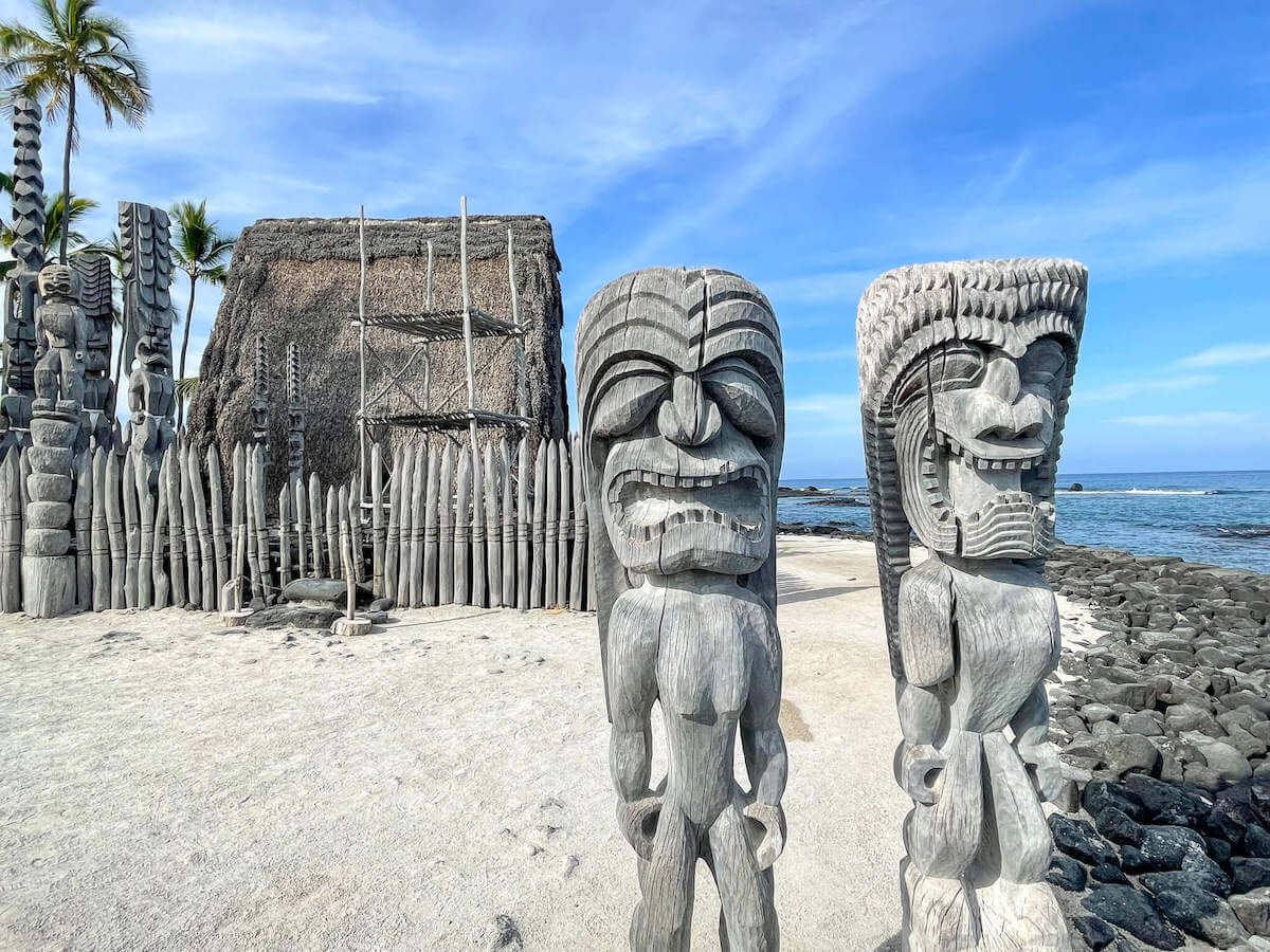 Find out how to explore the Hawaii Place of Refuge (also called Pu'uhonua o Honaunau) by top Hawaii blog Hawaii Travel with Kids. Image of two tikis with a Hawaiian hut and sand in the background.