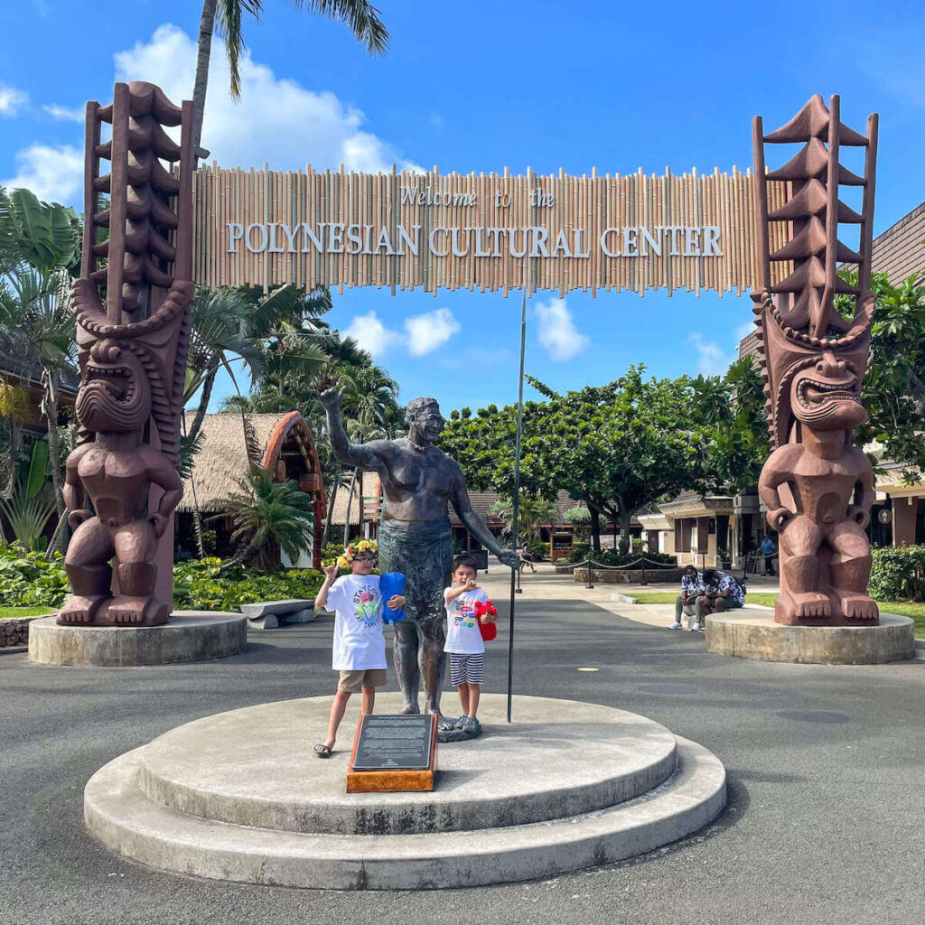 Read this full guide to the Polynesian Cultural Center in Hawai by top Hawaii blog Hawaii Travel with Kids. Image of two boys posing at the Polynesian Cultural Center entrance with tikis.