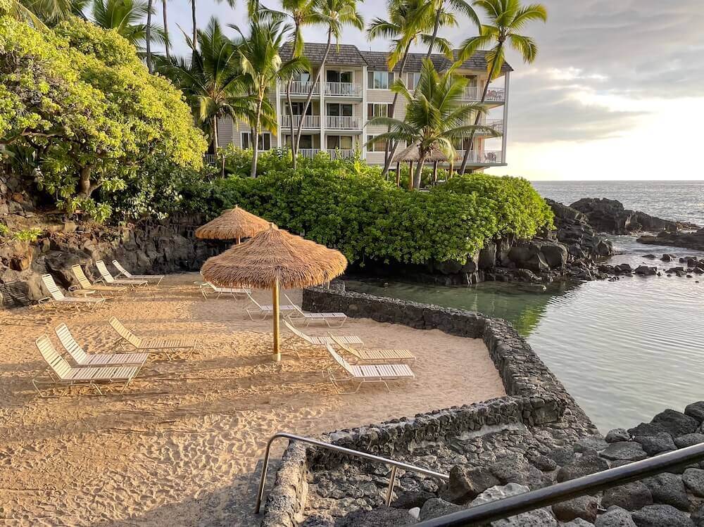 Image of a bunch of pool chairs on sand next to a lava rock wall and lagoon.