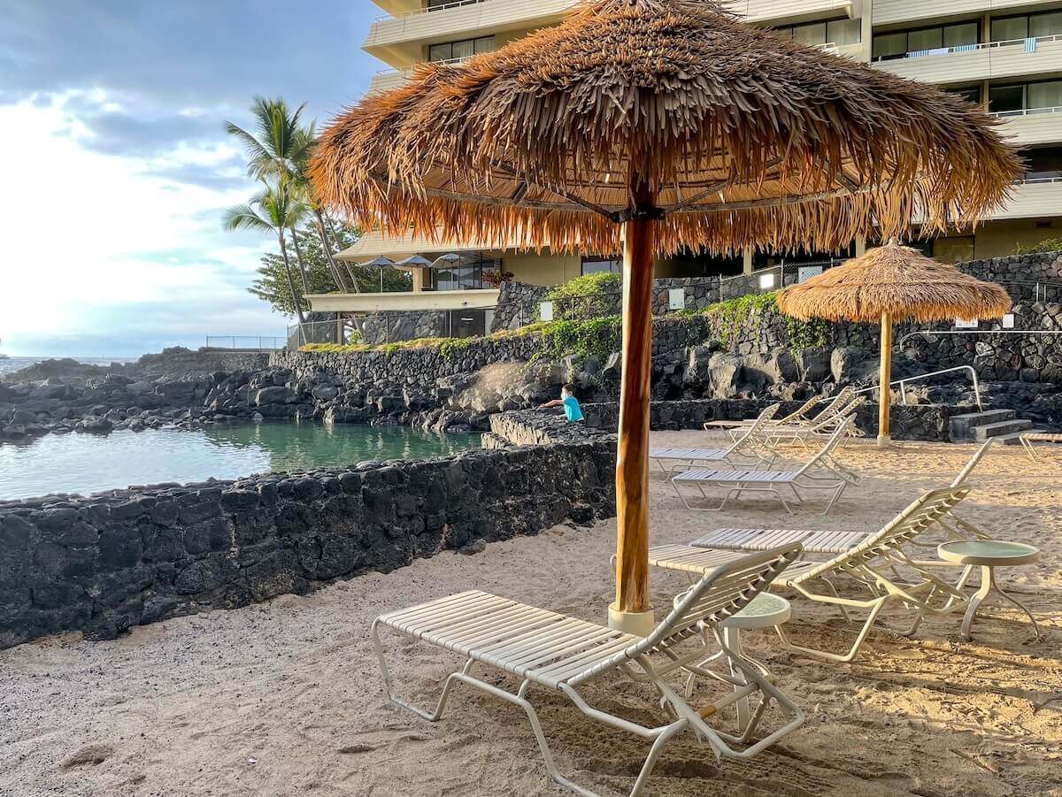 Read this honest Royal Kona Resort review by top Hawaii blog Hawaii Travel with Kids. Image of pool chairs under a thatched umbrella near the ocean.