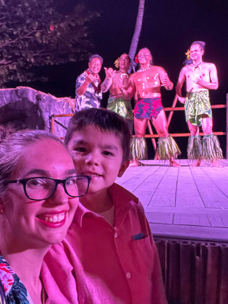 Image of a woman wearing glasses and a boy taking a selfie in front of male Polynesian dancers at a Big Island luau.