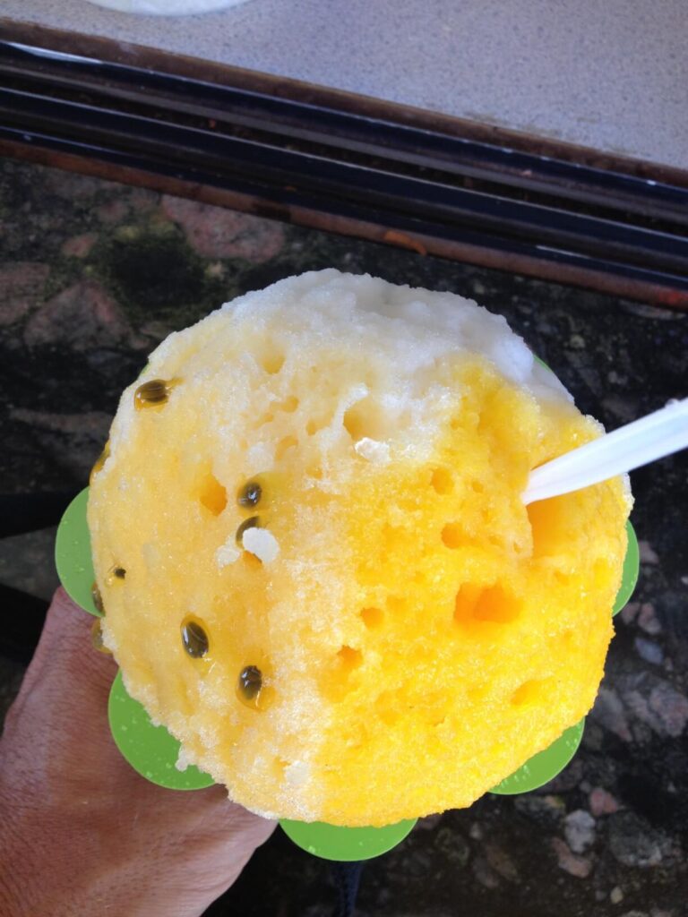 One of the best places for shave ice on Maui is Ululani's Shave Ice. Image of a cup of shave ice with lilikoi seeds and yellow syrup on top.
