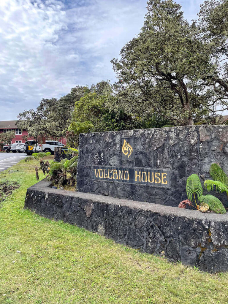 Image of the Volcano House sign on a lava rock wall with a red hotel in the background.