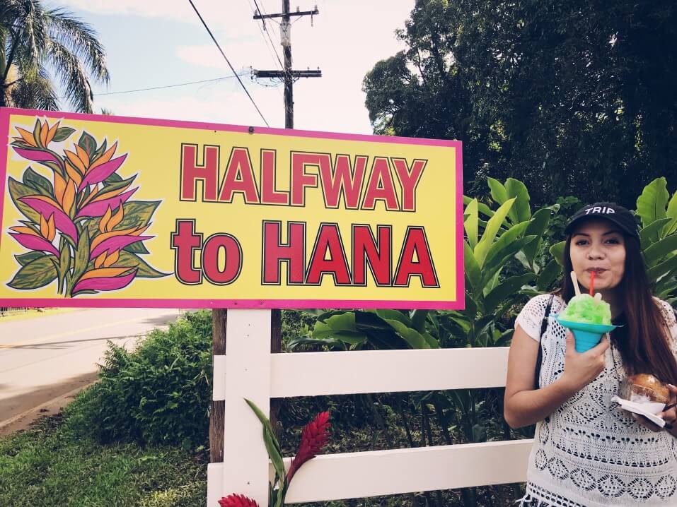 The Halfway to Hana roadside stand is a great place to get shave ice on Maui. Image of a woman eating shave ice next to the Halfway to Hana sign.