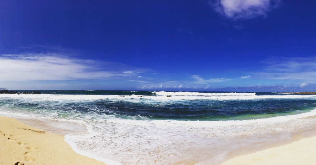 One of the best North Shore beaches Oahu offers is Pupukea Beach Park. Image of blue sky with clouds, blue water, and a white sandy beach.