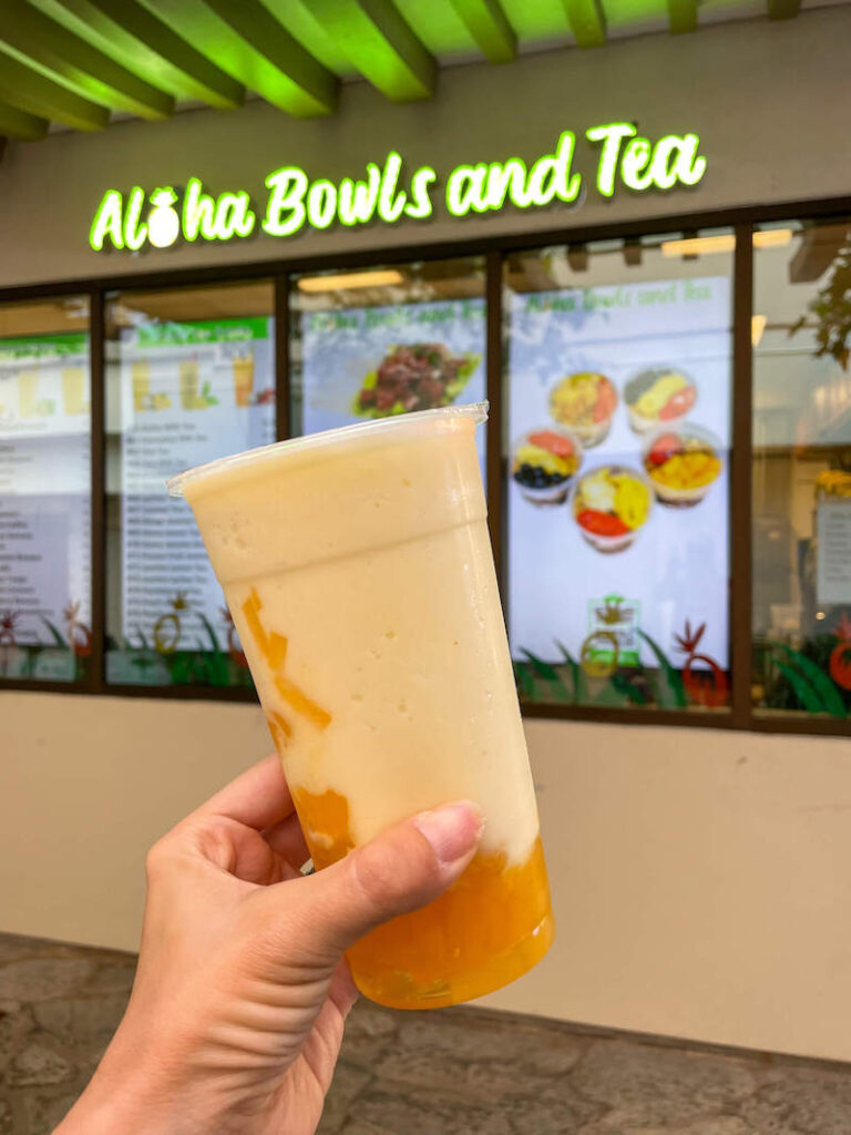 Image of a woman's hand holding up a mango bubble tea in front of the Aloha Bowls and Tea shop at the Hilton Hawaiian Village.