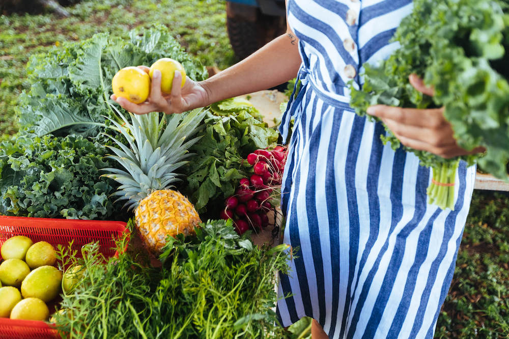 Find out the best Kauai farmers markets by top Hawaii blog Hawaii Travel with Kids. Image of a woman wearing a striped dress holding produce at a farmers market on Kauai.