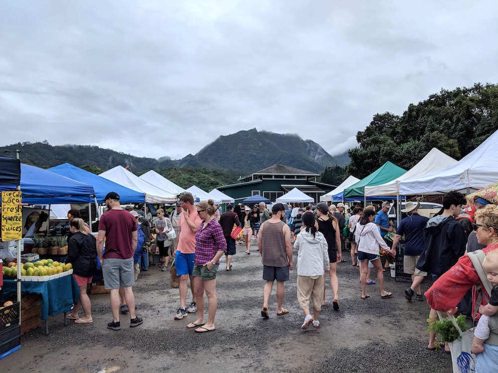 Image of people walking around a farmers market on Kauai on a cloudy day with mountains in the background.