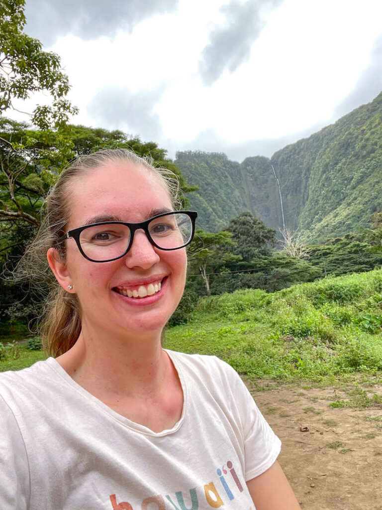 Image of a woman taking a selfie in the Waipio Valley with Hi'ilawe Falls in the background.